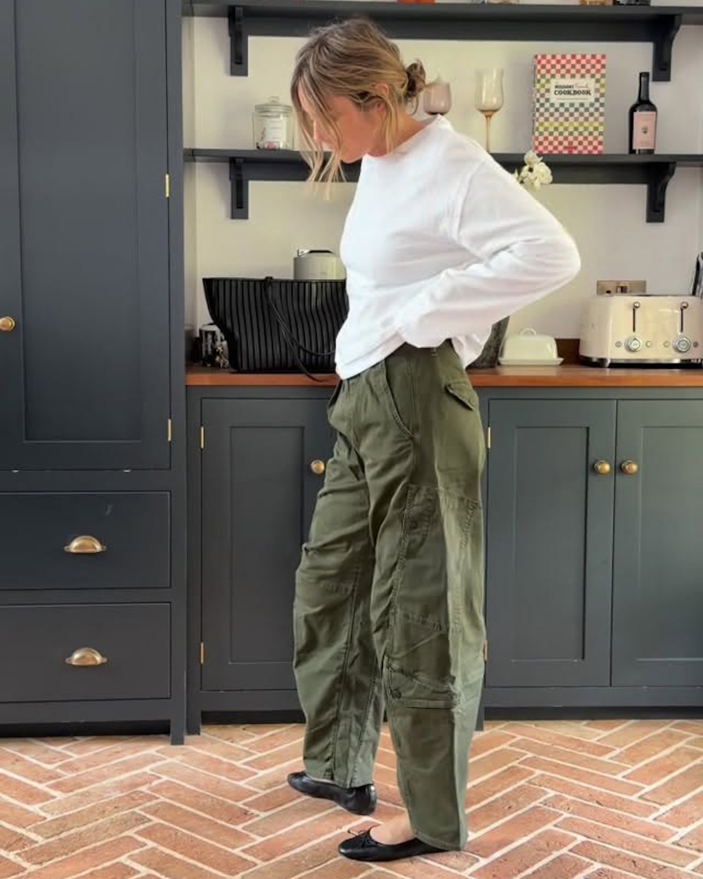 英國時尚達人艾瑪·羅斯·撒切爾 (Emma Rose Thatcher) 在她的廚房裡擺出造型，身穿超大號長袖白色 T 卹、黑色編織包、綠色筒褲和黑色芭蕾平底鞋。