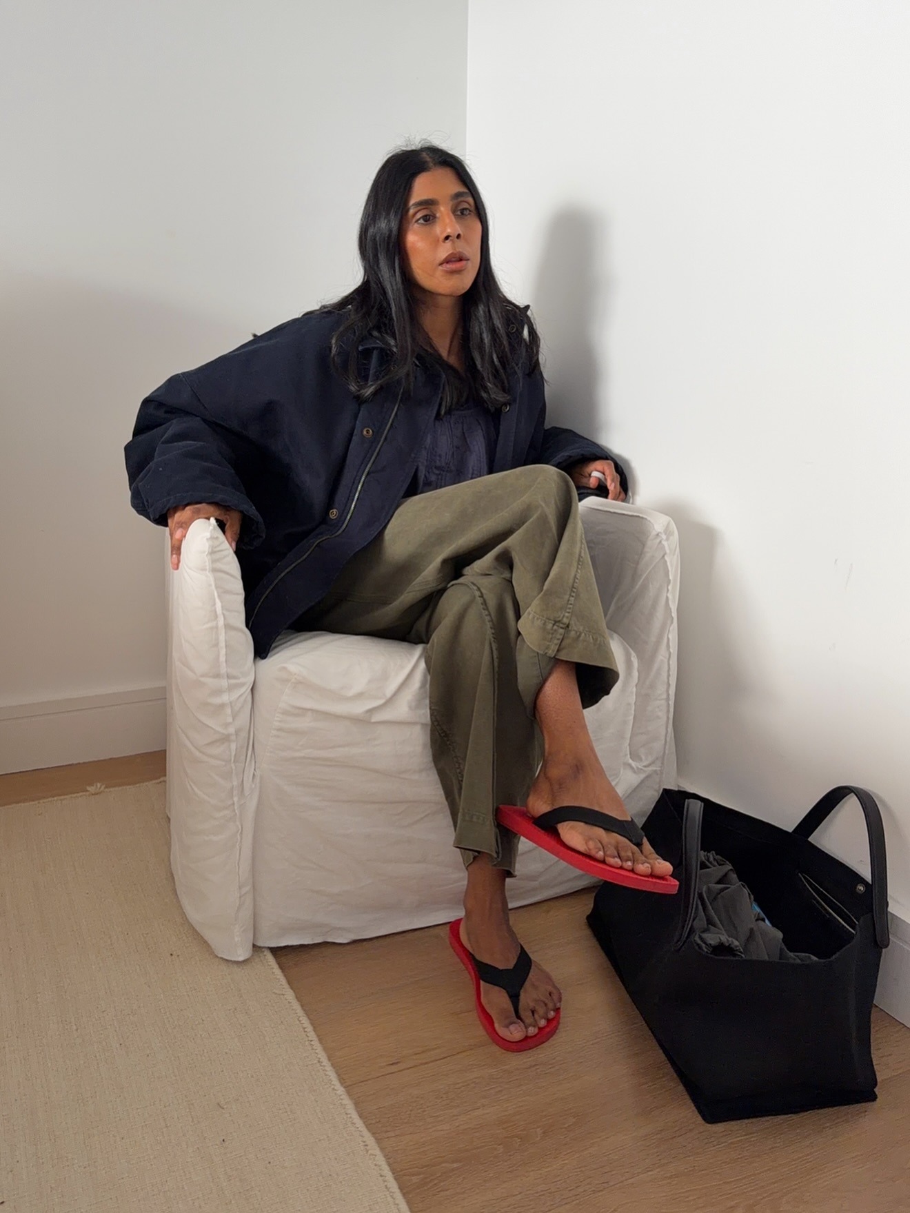英國時尚達人 Monikh Dale 坐在白色沙發椅上，身穿超大海軍藍色夾克、寬鬆綠色褲子、黑色絨面革手提包和黑色和紅色人字拖涼鞋。